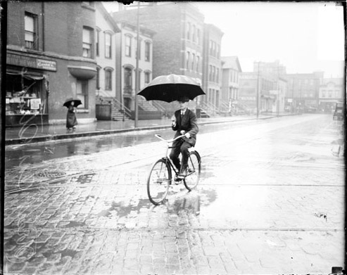 Image of a man holding an umbrella while riding a bike in the rain along an street during a street car strike in Chicago, Illinois. Street car tracks are visible on the street. View looking across the street with man at an angle to the camera. [ca. 1915 June 15] Chicago Daily News negatives collection, DN-064587. Courtesy of Chicago History Museum.