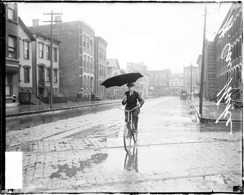 Image of a man holding an umbrella while riding a bike along an street in the rain during a street car strike in Chicago, Illinois. Street car tracks are visible on the street. View looking down the street with the man facing the camera. [ca. 1915 June 15] Chicago Daily News negatives collection, DN-064588. Courtesy of Chicago History Museum.